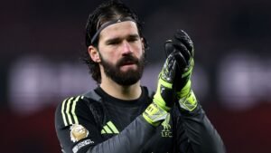 Liverpool-doelman Alisson Becker applaudisseert voor de fans, gekleed in een zwart tenue met groene accenten en handschoenen, terwijl hij zijn gefocuste uitdrukking toont.
