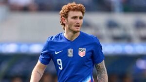 L'attaquant de l'USMNT Josh Sargent en maillot bleu, numéro 9, se concentre intensément pendant un match, mettant en valeur ses cheveux bouclés caractéristiques.