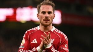 يبدو ماك أليستر من ليفربول مفكراً وهو يرتدي الزي الأحمر للفريق، يصفق للجماهير بعد المباراة مع خلفية ضبابية للحشد.