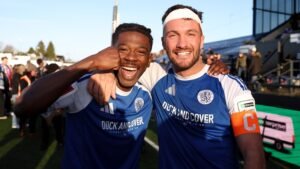 Macclesfield-spelers vieren vrolijk, gekleed in blauwe shirts, na een wedstrijdoverwinning, waarbij één een aanvoerdersband toont, met fans op de achtergrond.