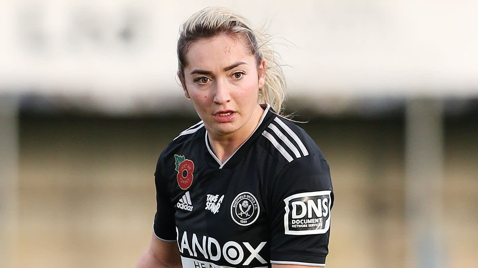 Maddy Cusack portant le maillot noir de Sheffield United avec l'emblème du coquelicot, concentrée sur le terrain pendant un match, faisant preuve de détermination.