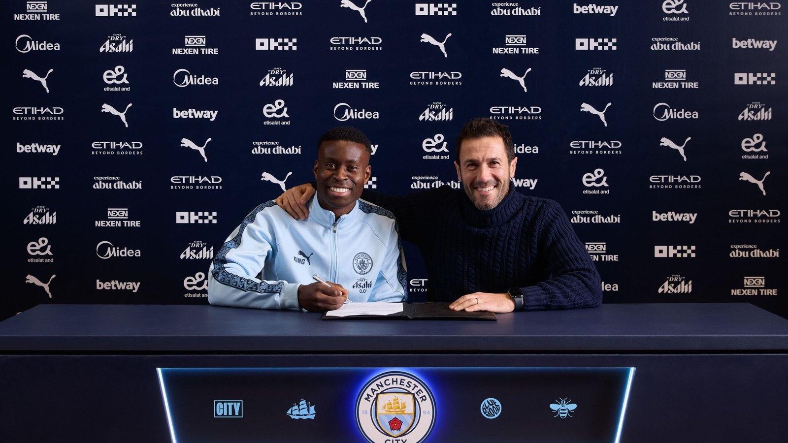 Marc Guehi sourit en signant avec Manchester City aux côtés d'un représentant, tous deux assis à une table ornée des logos du club.