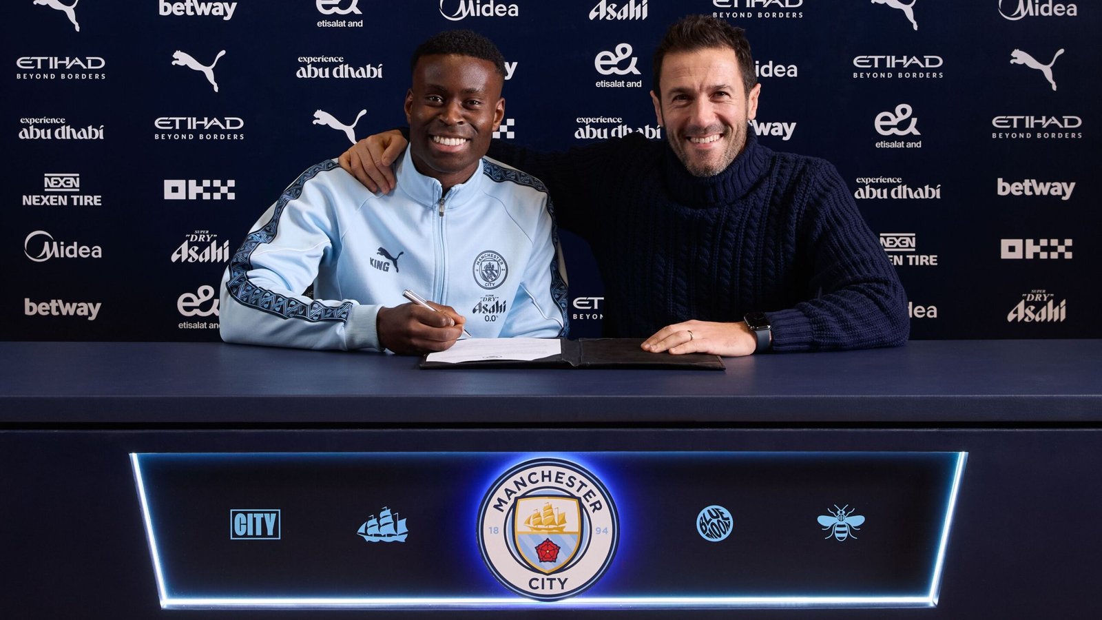 Marc Guehi signe son contrat avec Manchester City, souriant aux côtés d'un représentant du club à la table de signature, avec l'identité visuelle du club.