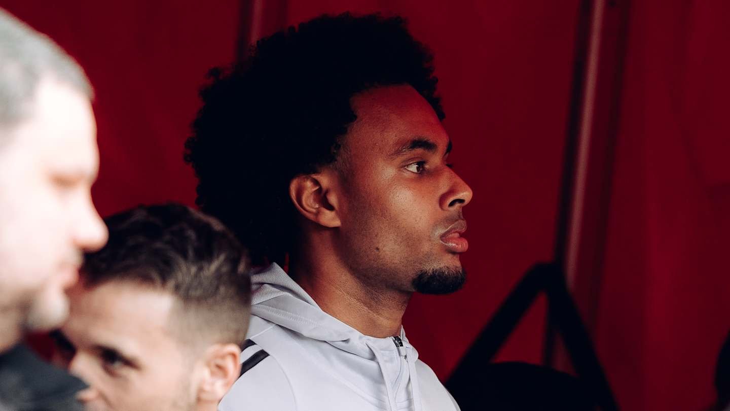 Joshua Zirkzee en profil sous un fond rouge, dévoilant ses cheveux bouclés et son expression concentrée, au milieu de coéquipiers flous.