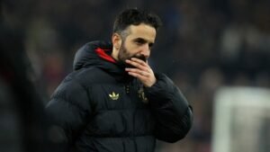 Amorim, dressed in a black Manchester United jacket, contemplates with a thoughtful expression, amid a blurred stadium background.