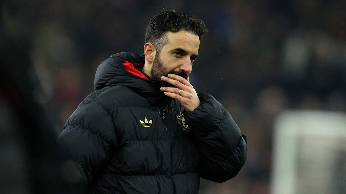 Amorim, dressed in a black Manchester United jacket, contemplates with a thoughtful expression, amid a blurred stadium background.