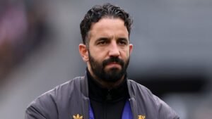 مدرب كرة قدم مغربي بلحية كثيفة، يرتدي سترة سوداء ذات سحاب، يحدق بتأمل على خلفية ملعب ضبابية.