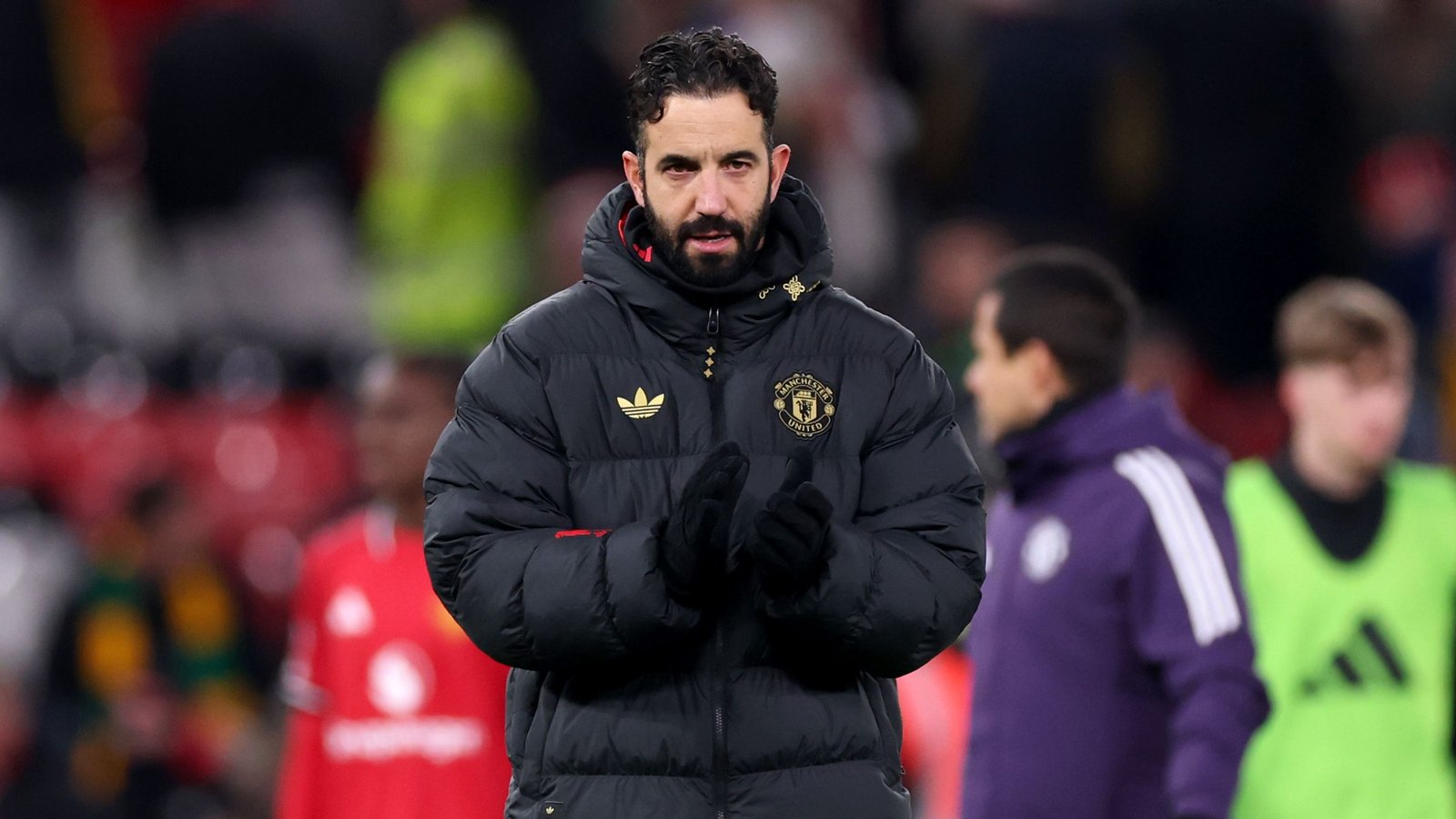 L'entraîneur de Manchester United, vêtu d'une doudoune noire arborant l'emblème du club, applaudit dans un stade entouré de fans et de joueurs flous.