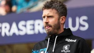 Michael Carrick staat peinzend aan de zijlijn, gekleed in een zwarte hoodiejas met logo's, tegen een wazige stadionachtergrond.