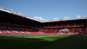 Open ruimte op Old Trafford, met zicht op de Sir Alex Ferguson-tribune en levendige rode stoelen onder een helderblauwe hemel, klaar voor de wedstrijd.