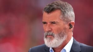 Roy Keane, in een donker pak en wit overhemd, is te zien met een geconcentreerde uitdrukking, tegen een wazige rode achtergrond.