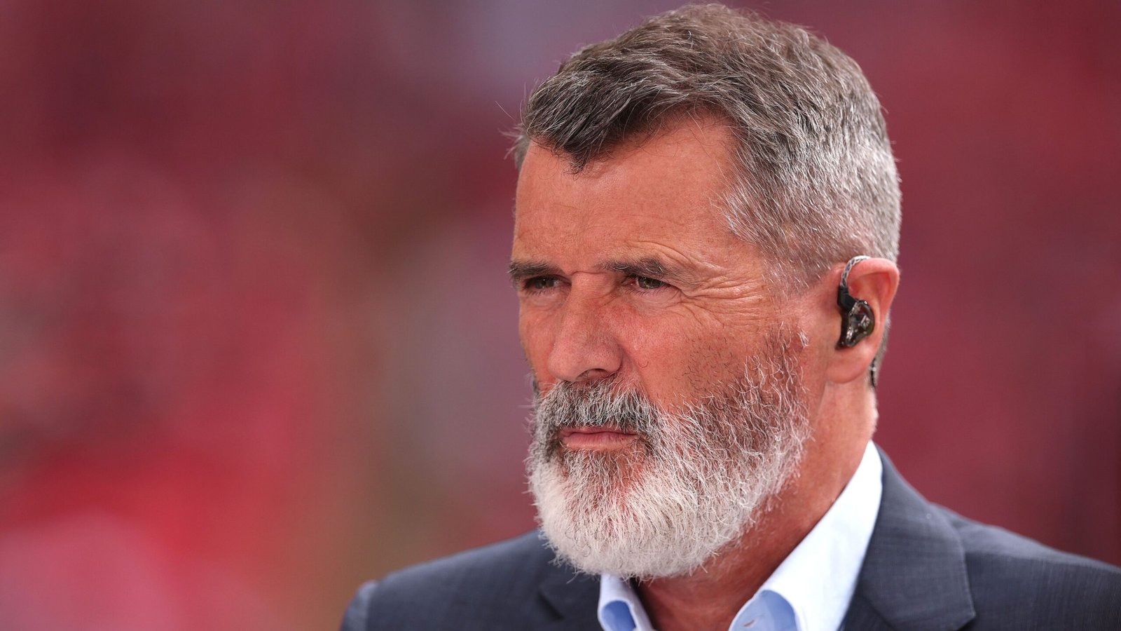 Roy Keane, in een donker pak en wit overhemd, is te zien met een geconcentreerde uitdrukking, tegen een wazige rode achtergrond.