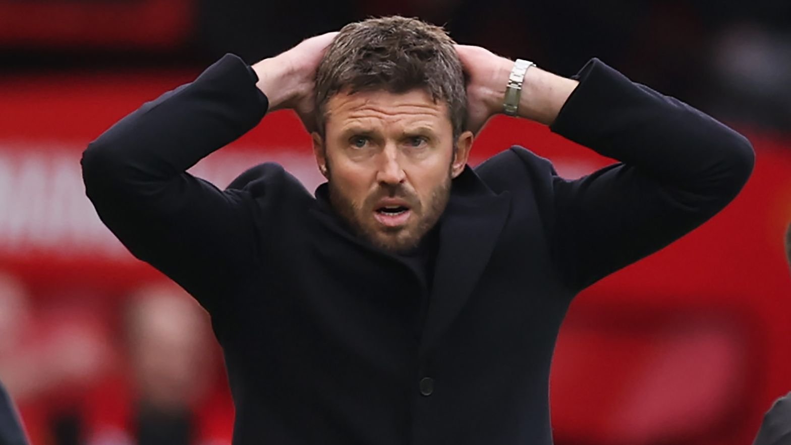 Een bezorgde Manchester United-coach die reageert tijdens een gespannen moment, met handen op het hoofd en een zwarte jas, tegen een levendige rode achtergrond.