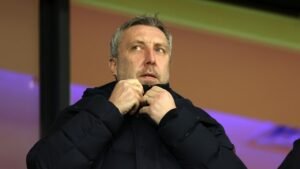 Serieus uitziende voetbalmanager past zijn jas aan, met een vervaagde kleurrijke achtergrond die de sfeer van een wedstrijd vastlegt.