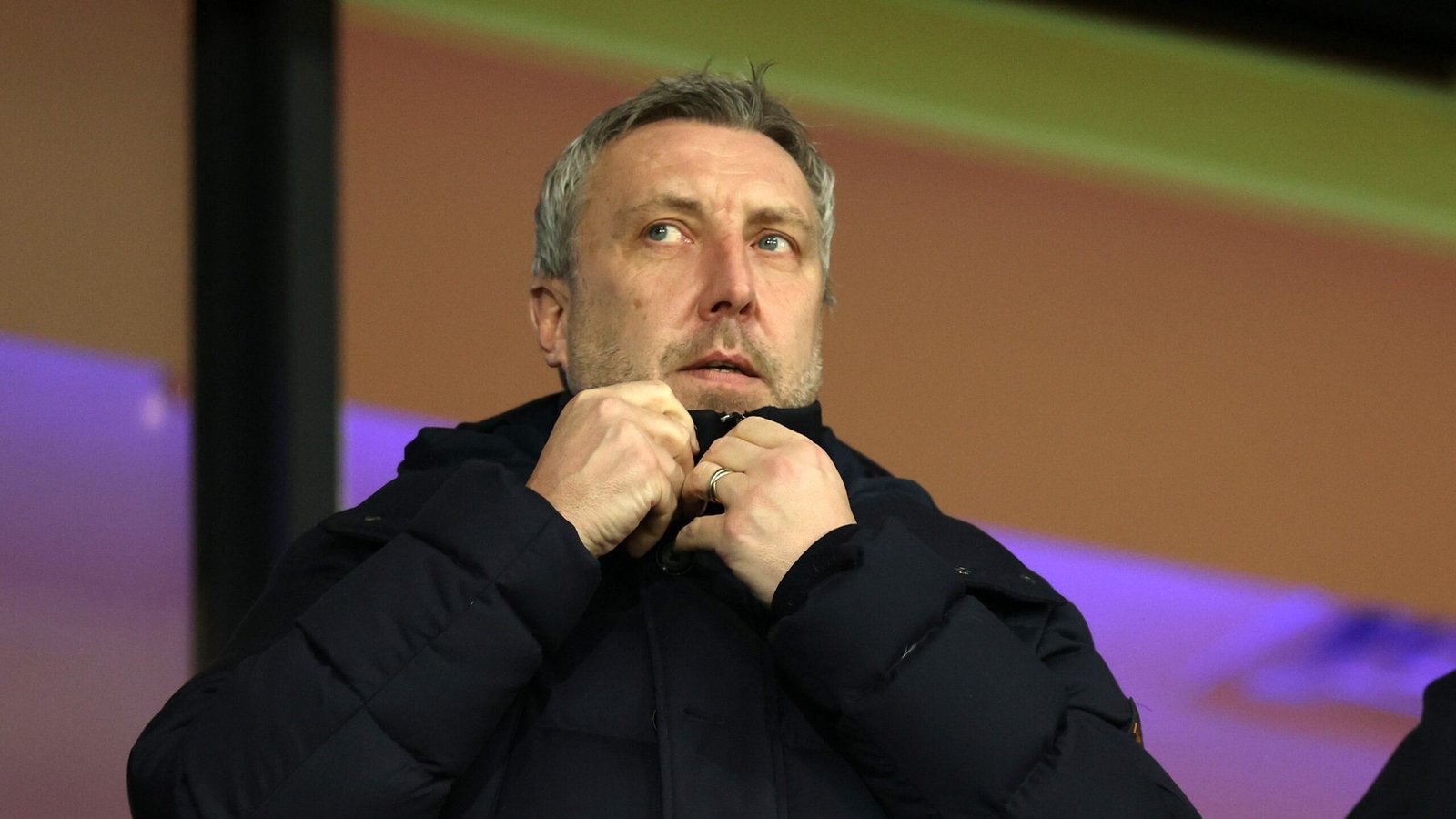 Serieus uitziende voetbalmanager past zijn jas aan, met een vervaagde kleurrijke achtergrond die de sfeer van een wedstrijd vastlegt.