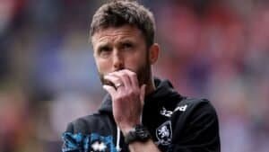 Michael Carrick réfléchit pendant un match, vêtu d'un haut d'entraînement noir avec des accents bleus, illustrant son approche stratégique du coaching.