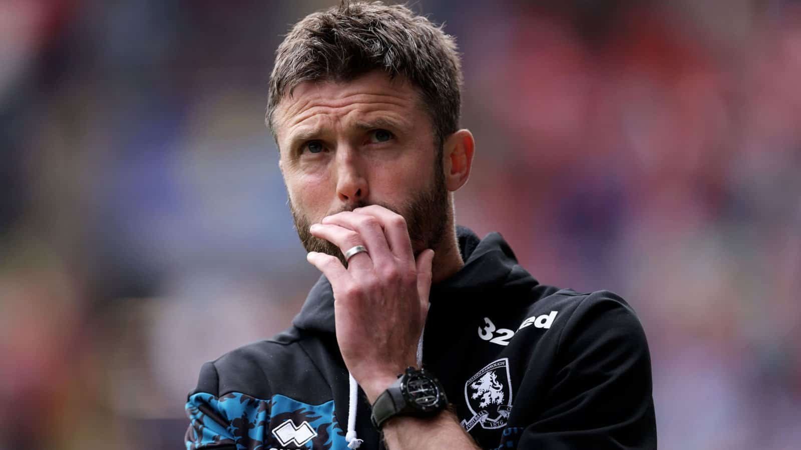 Michael Carrick réfléchit pendant un match, vêtu d'un haut d'entraînement noir avec des accents bleus, illustrant son approche stratégique du coaching.