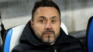 Roberto De Zerbi, gekleed in een zwart jasje, staart peinzend vanaf de bank tijdens een wedstrijd, wat zijn focus en intensiteit benadrukt.