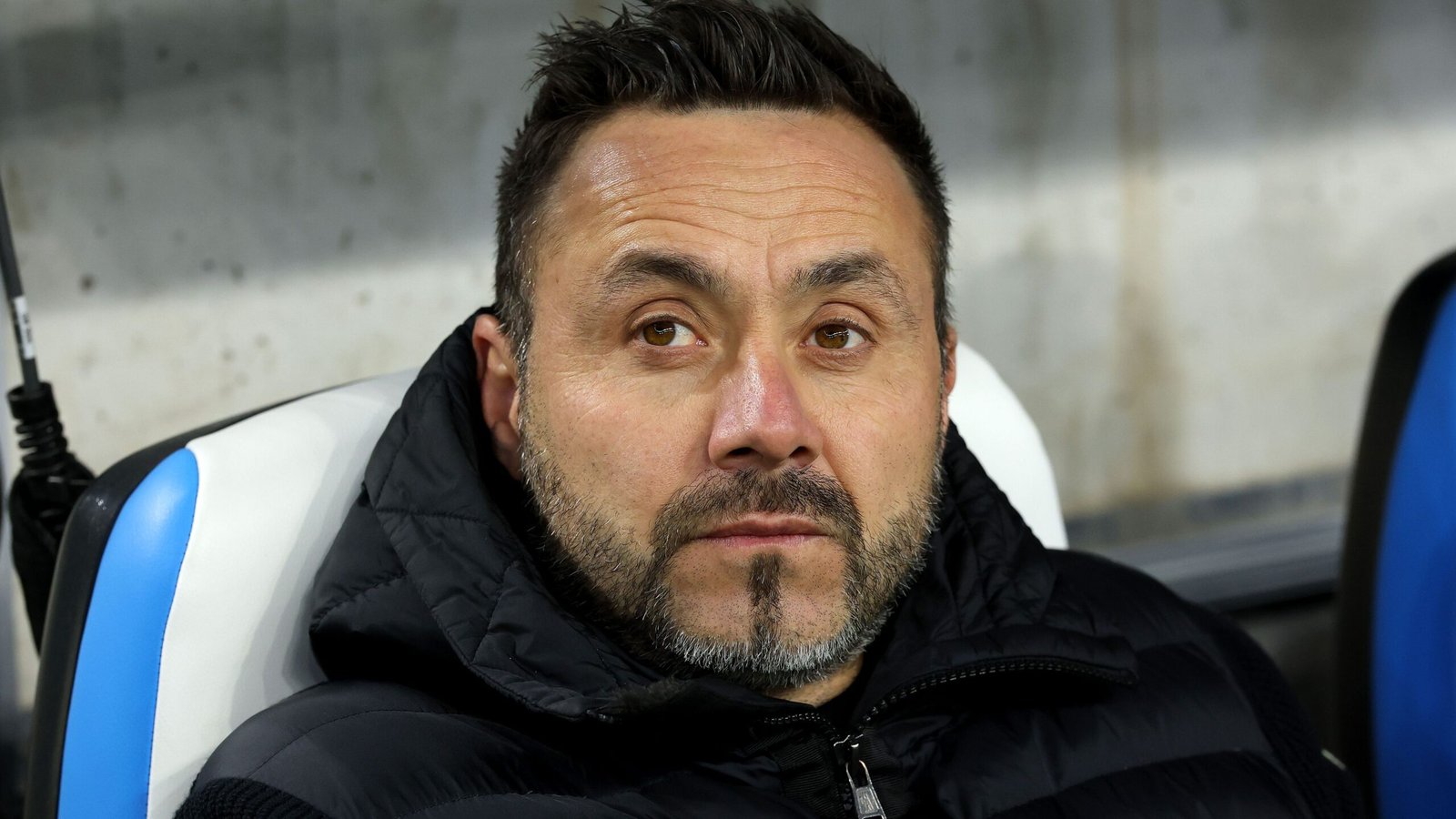 Roberto De Zerbi, gekleed in een zwart jasje, staart peinzend vanaf de bank tijdens een wedstrijd, wat zijn focus en intensiteit benadrukt.