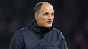 Thomas Tuchel glimlacht tijdens een wedstrijd, hij draagt een donker jasje tegen een wazig stadionachtergrond, wat zijn focus op voetbalstrategie benadrukt.