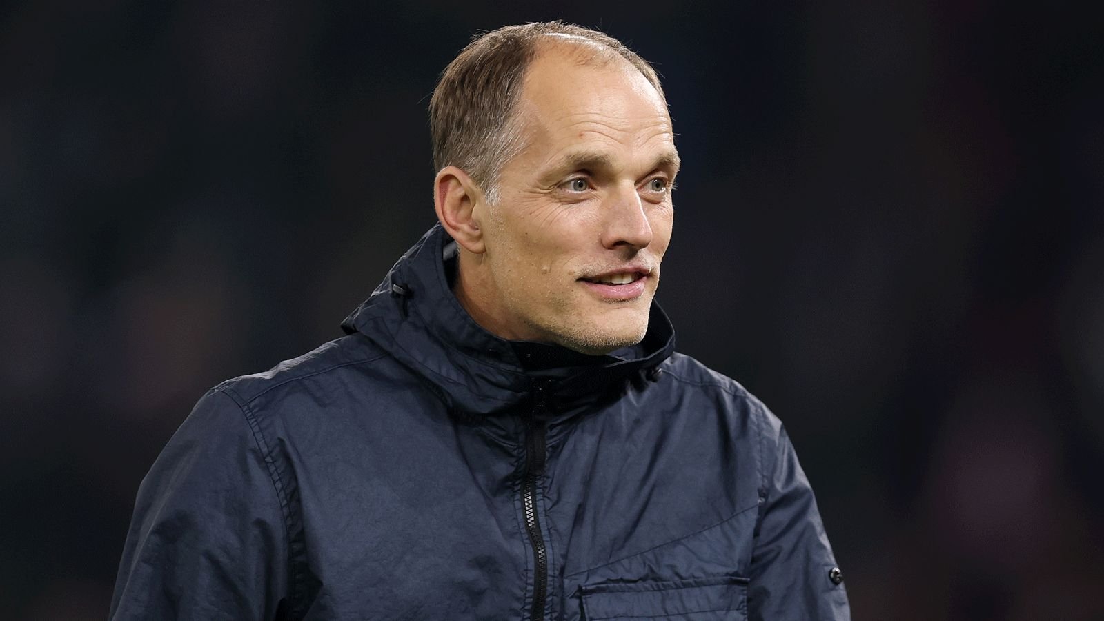 Thomas Tuchel glimlacht tijdens een wedstrijd, hij draagt een donker jasje tegen een wazig stadionachtergrond, wat zijn focus op voetbalstrategie benadrukt.