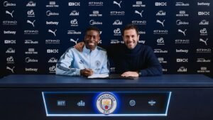 Marc Guehi sourit alors qu'il signe avec Manchester City, assis aux côtés d'un représentant du club devant un fond orné des logos de l'équipe.