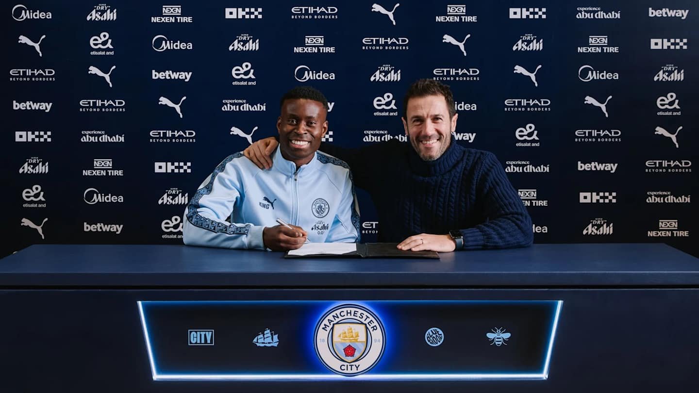 Marc Guehi sourit alors qu'il signe avec Manchester City, assis aux côtés d'un représentant du club devant un fond orné des logos de l'équipe.