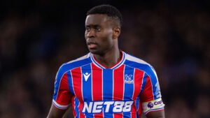 Marc Guehi, portant le maillot rayé rouge et bleu de Crystal Palace avec le logo du sponsor 'NET88', se concentre intensément pendant un match.