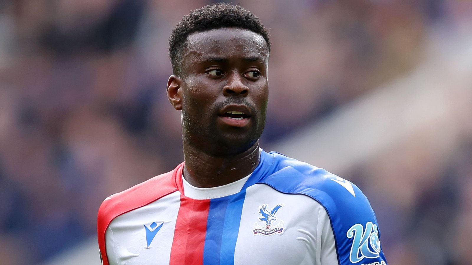 Marc Guehi, portant le maillot rouge et bleu de Crystal Palace, concentré pendant un match, faisant preuve de détermination sur le terrain.