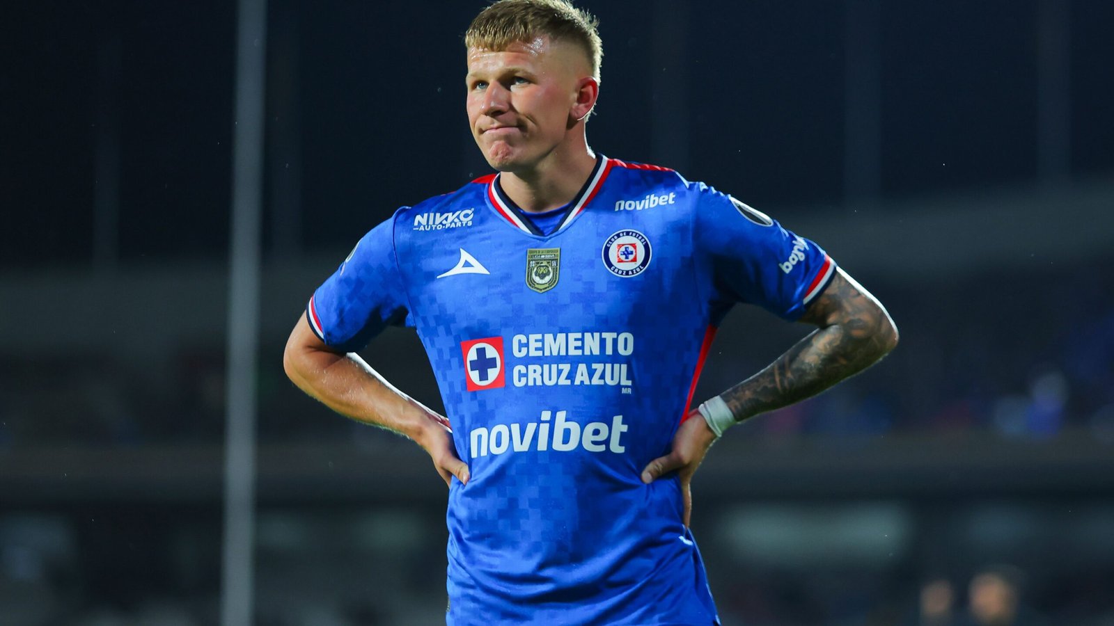 Mateusz Bogusz in een blauw Cruz Azul-tenue, peinzend na een wedstrijd, met stadionlichten op de achtergrond die de spanning benadrukken.