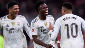 Mbappé célèbre avec Vinícius et Jude Bellingham, tous en tenue blanche du Real Madrid, exprimant la joie après un moment de match.