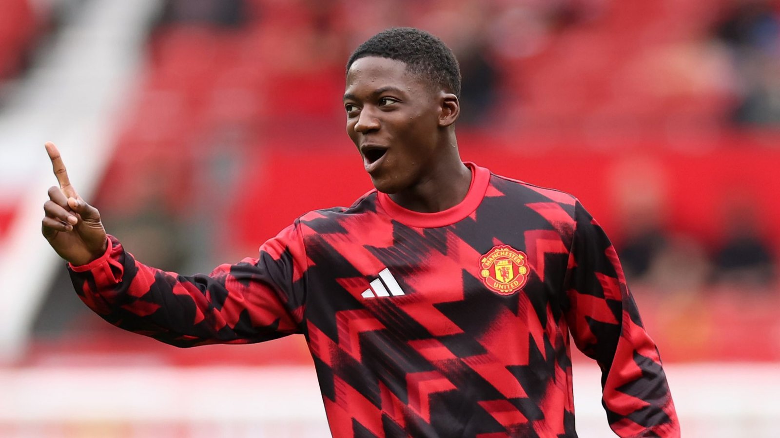 كوبي ماينو يحتفل، مرتدياً طقم التدريب الأحمر والأسود لمانشستر يونايتد، مع أجواء الملعب النابضة بالحياة في الخلفية.
