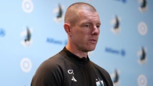 Cam Knowles, hoofdtrainer van Minnesota United, spreekt op een persconferentie, met een blauwe achtergrond met clublogo's en afbeeldingen.
