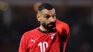 Mohamed Salah draagt een rood Egypte-tenue, kijkt peinzend met zijn hand op zijn hoofd tijdens een wedstrijd, wat zijn emotionele reactie na het spel toont.
