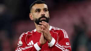 محمد صلاح، مرتديًا قميص ليفربول الأحمر، يصفق بتعبير مركز، مُظهرًا الشغف والتفاني وسط أجواء المباراة.