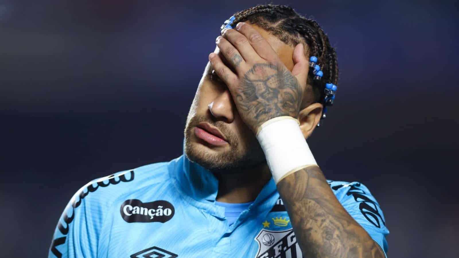 Neymar in een blauw voetbaltenue, zichtbaar aangedaan, met zijn hand op zijn gezicht, waarbij zijn getatoeëerde armen en gevlochten haar te zien zijn.