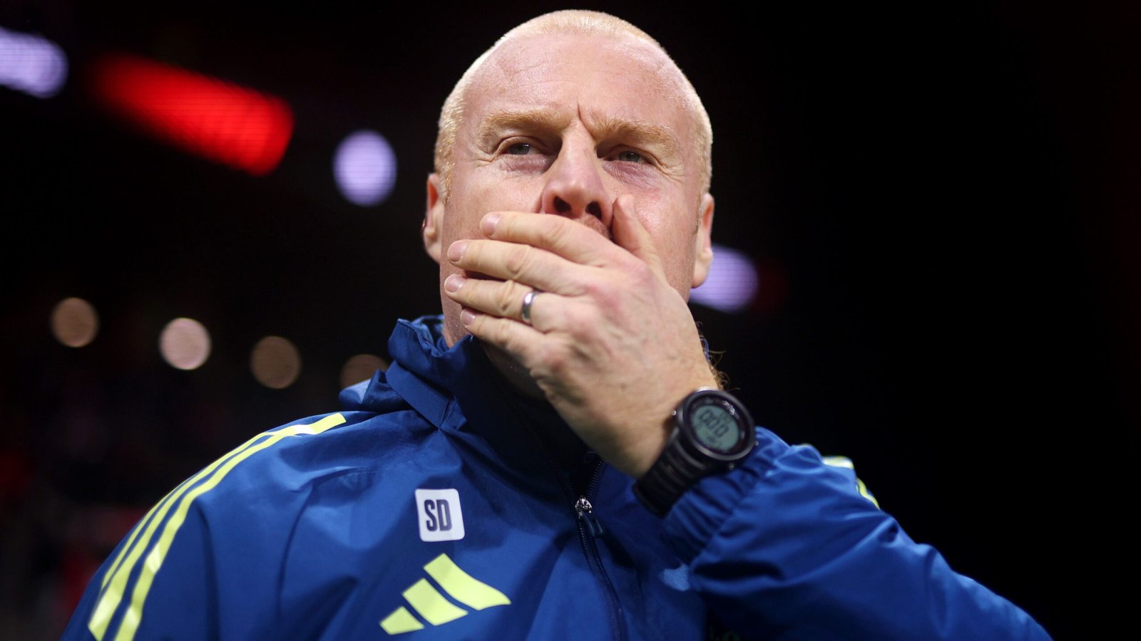 Een geconcentreerde coach in een blauw trainingspak bedekt zijn mond en toont intense concentratie en bezorgdheid tijdens een gespannen moment in een wedstrijd.