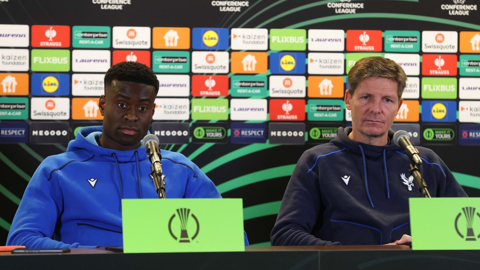 De manager en een speler van Crystal Palace zitten op een persconferentie, beide in blauwe hoodies, tegen een achtergrond van levendige sponsorlogo's voor de UEFA Conference League.