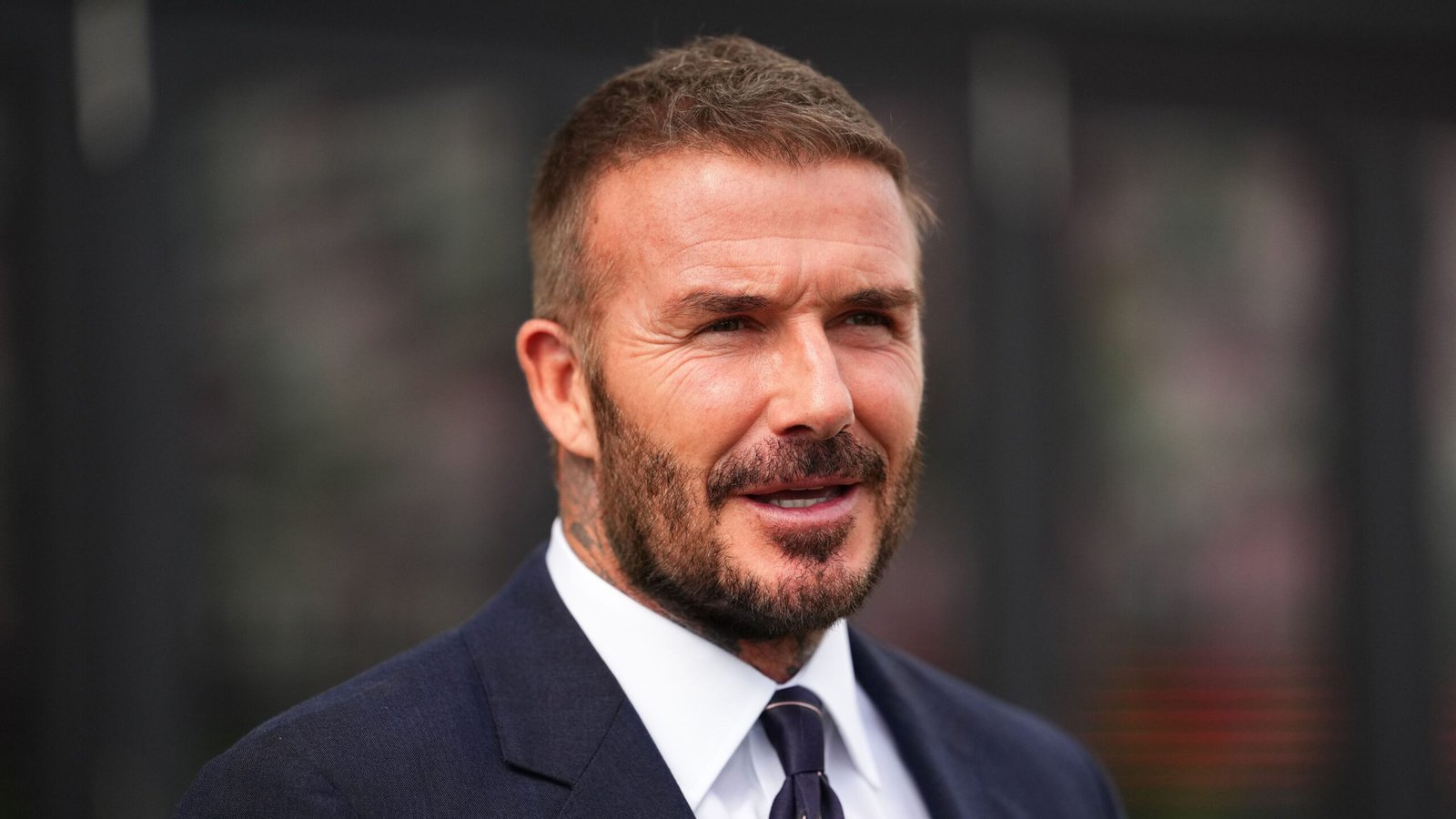 David Beckham sourit, portant un costume sombre et une cravate, avec un arrière-plan flou évoquant une apparition publique.