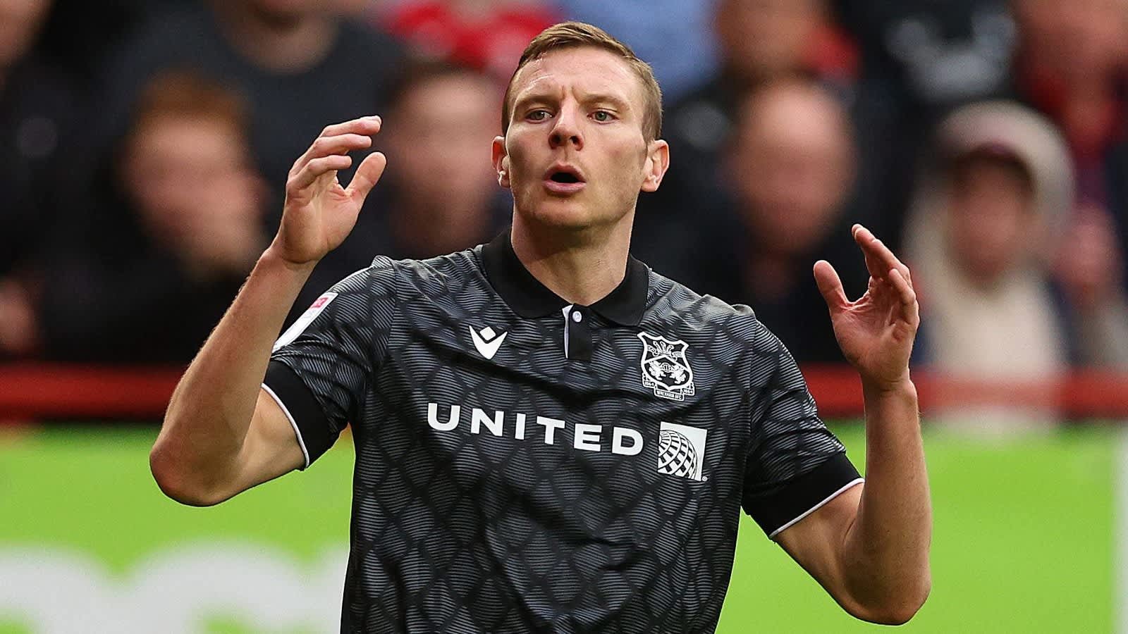 Paul Mullin gesticule sur le terrain, portant le maillot extérieur noir de Wrexham avec le logo "United", lors d'un moment intense du match.