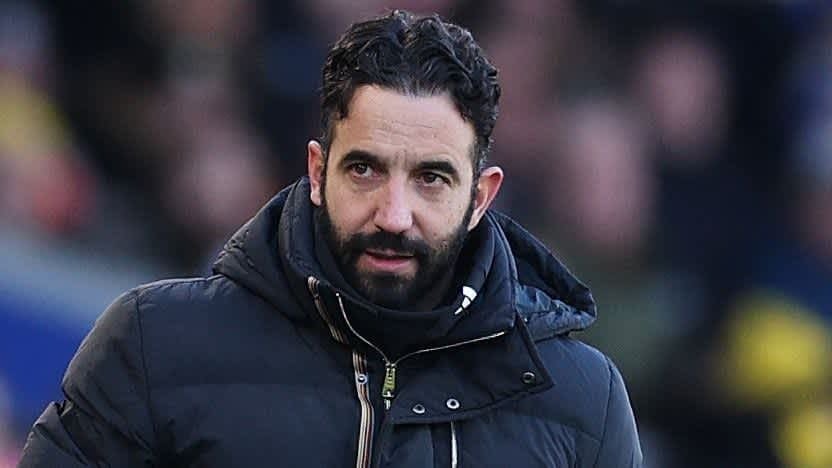 Rúben Amorim, gekleed in een zwarte gevoerde jas, kijkt bedachtzaam tijdens een voetbalwedstrijd, met wazige fans op de achtergrond.