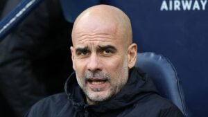 Pep Guardiola toont een geconcentreerde uitdrukking terwijl hij op de bank zit, gekleed in een zwart jasje, tegen een blauwe achtergrond.