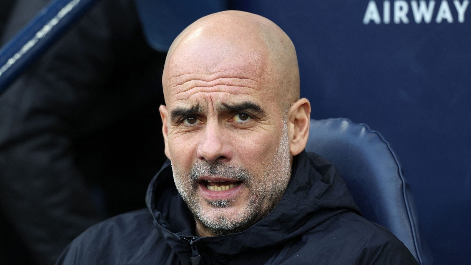 Pep Guardiola toont een geconcentreerde uitdrukking terwijl hij op de bank zit, gekleed in een zwart jasje, tegen een blauwe achtergrond.