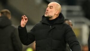 Pep Guardiola gebaart expressief tijdens een wedstrijd, gekleed in een zwart jasje, tegen de achtergrond van een stadionpubliek.