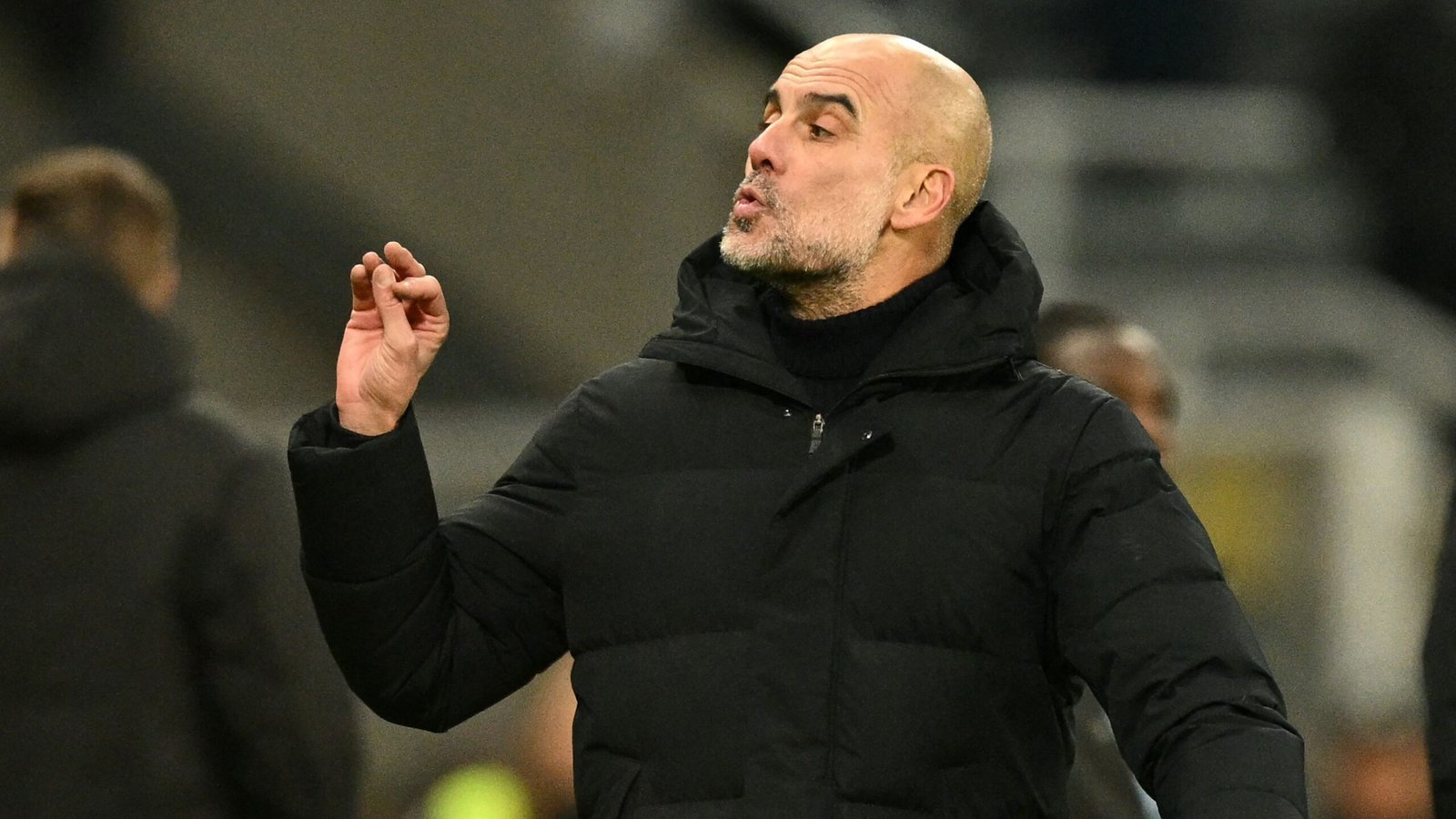 Pep Guardiola gebaart expressief tijdens een wedstrijd, gekleed in een zwart jasje, tegen de achtergrond van een stadionpubliek.