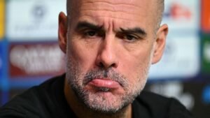 Pep Guardiola oogt serieus tijdens een persconferentie, met een gefronst voorhoofd en een subtiele frons, terwijl op de achtergrond vervaagde teamlogo's te zien zijn.