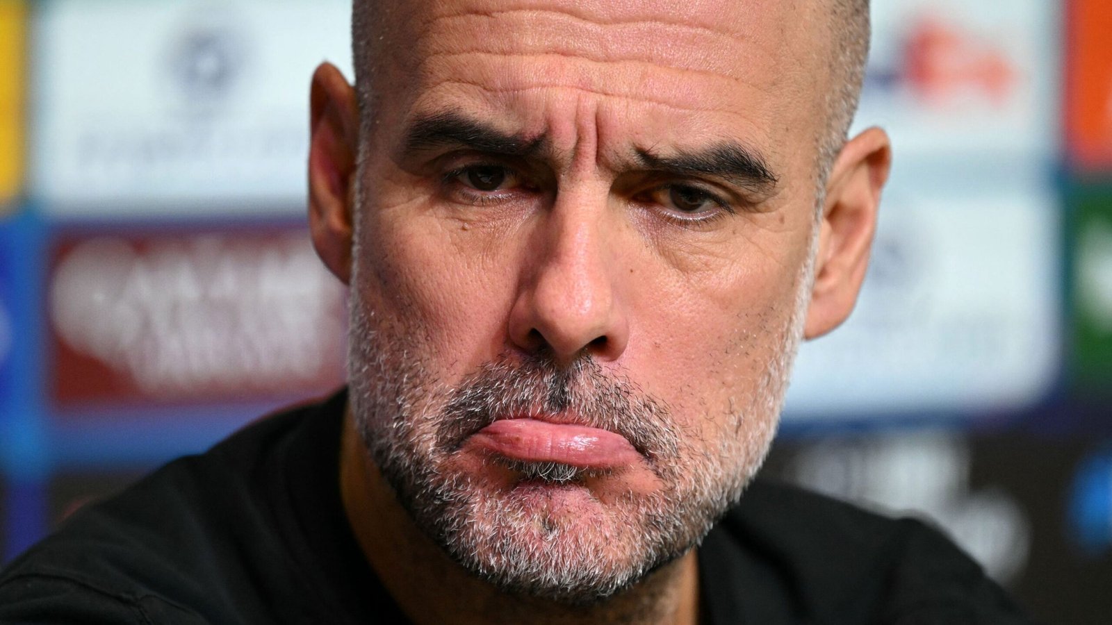 Pep Guardiola oogt serieus tijdens een persconferentie, met een gefronst voorhoofd en een subtiele frons, terwijl op de achtergrond vervaagde teamlogo's te zien zijn.