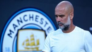 Pep Guardiola, gekleed in een wit shirt, kijkt peinzend naar beneden voor het logo van Manchester City, waarbij de kleuren van de club worden getoond.