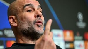 Pep Guardiola gebaart tijdens een persconferentie van Manchester City, wat een serieuze discussie benadrukt in een donkere setting met sponsorlogo's op de achtergrond.