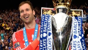 Petr Čech célèbre avec le trophée de Premier League, vêtu de sa tenue de gardien de but, au milieu d'une foule en liesse. Des rubans bleus et blancs ornent le trophée.